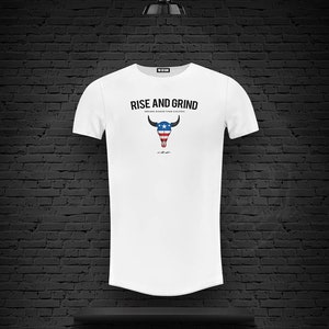 Puede incluir: Camiseta blanca de cuello redondo y mangas cortas. La camiseta tiene un estampado gráfico de un cráneo de toro con un diseño de bandera estadounidense. El texto "Rise and Grind" está impreso sobre el gráfico. El texto "Dreams bigger than excuses" está impreso debajo del gráfico.