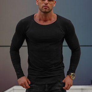 Puede incluir: Un hombre con una camisa negra de manga larga y pantalones vaqueros de color oscuro. Lleva gafas de sol y un reloj de oro.