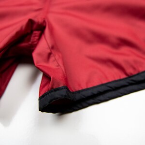 K&ouml;nnte beinhalten: Nahaufnahme von roten Sporthosen mit schwarzem Besatz. Die Shorts bestehen aus einem glatten Stoff, m&ouml;glicherweise Nylon oder Polyester. Der schwarze Besatz umrandet den unteren Saum der Shorts.