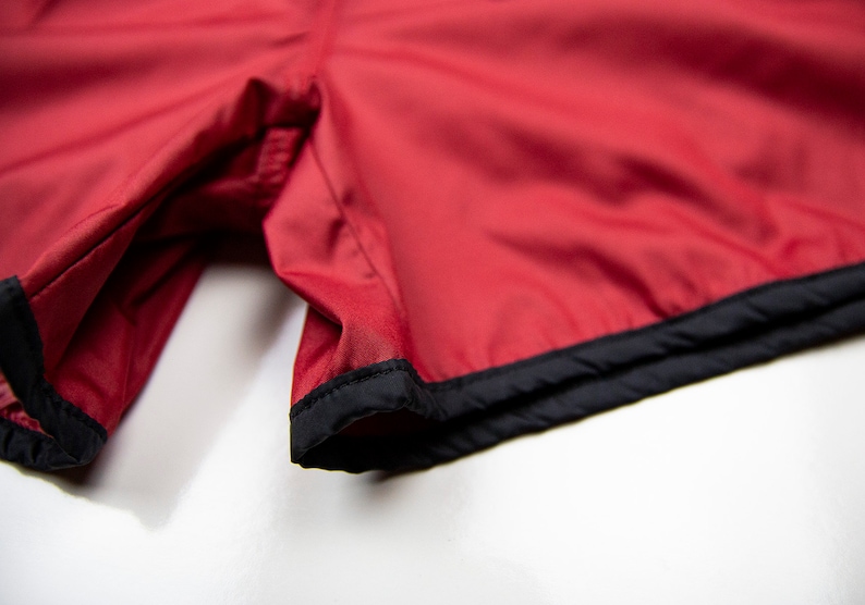 K&ouml;nnte beinhalten: Nahaufnahme von roten Sporthosen mit schwarzem Besatz. Die Shorts bestehen aus einem glatten Stoff, m&ouml;glicherweise Nylon oder Polyester. Der schwarze Besatz umrandet den unteren Saum der Shorts.