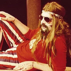 May include: A man with long blonde hair and a beard is wearing a red shirt, striped pants, and sunglasses. He is lying down with his legs crossed and his arms resting on his lap.