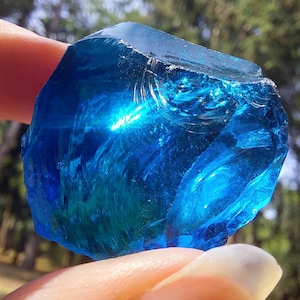 Può includere: Un grande cristallo blu sfaccettato con una superficie liscia e vetrosa. Il cristallo è tenuto in una mano, e lo sfondo è sfocato.