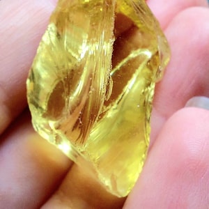 Peut inclure: Une roche de verre translucide jaune-orangé avec une surface rugueuse et texturée. La roche est tenue dans une main, avec une feuille verte floue en arrière-plan. La forme de la roche est irrégulière et angulaire.