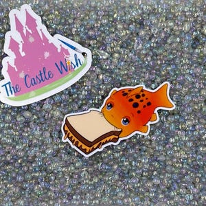May include: Two colorful stickers on a background of iridescent beads. One sticker features a pink castle with the text "The Castle Wish". The other sticker features a red and orange fish with black spots eating a peanut butter and jelly sandwich.