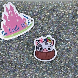 May include: A close-up of iridescent white and clear glass beads. Two stickers are on top of the beads. One sticker is pink and features a castle with the text "The Castle Wish". The other sticker is white and features a cartoon pig with a flower crown and a brown coconut shell.