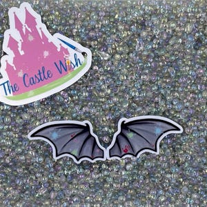 May include: A close-up of iridescent white glass beads with two black bat wing stickers on top. The stickers have a white paint splattered design. The sticker on the left has the text "The Castle Wish" in white.