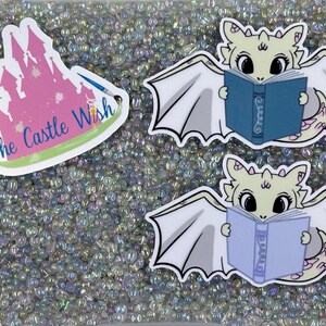 May include: Two white dragon stickers with wings, reading books. The dragon on the top is reading a blue book with the word "Story" on the cover. The dragon on the bottom is reading a purple book with the word "Dreams" on the cover. The stickers are on a background of iridescent beads.
