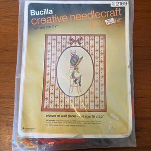May include: A Bucilla creative needlecraft kit in a clear plastic bag. The kit features a design of a person sitting on a chair, with the text "SITTING PRETTY". The cut size is 18 x 22 inches.