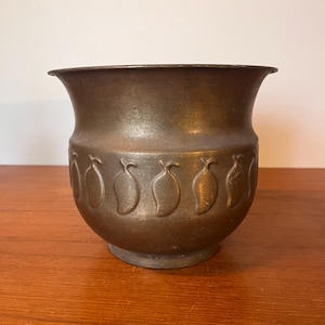 May include: A bronze metal planter with a flared rim and a rounded body. The planter has a repeating pattern of embossed fruit shapes around its midsection. It sits on a small, circular base. A decorative item for the home.
