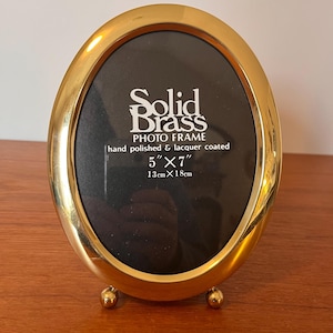 May include: Oval gold-colored photo frame with a black interior. The frame is supported by two small, round feet. Text on the frame reads "Solid Brass PHOTO FRAME hand polished & lacquer coated 5" x 7".