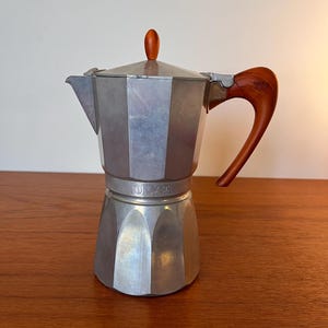 Op de afbeelding: Een zilveren espressomaker voor op het fornuis met een houten handvat en knop. De koffiepot heeft een klassiek ontwerp met een facetgeslepen body en een tuit om uit te schenken. De handgreep is gebogen en de knop is donkerbruin.