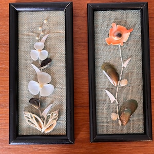 Peut inclure: Deux tableaux muraux encadrés sur le thème floral. La pièce de gauche présente une fleur blanche faite de coquillages sur un fond de toile de jute verte. La pièce de droite présente une fleur rouge faite de coquillages sur un fond de toile de jute verte.