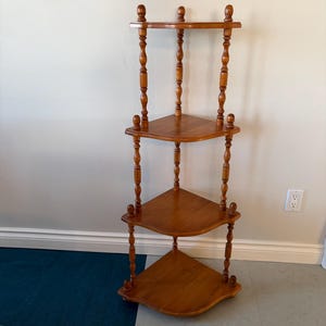May include: A wooden corner shelf with three triangular shelves. The shelves and supports are a warm, medium brown color. The supports have a turned spindle design, and the top of the shelf has decorative finials.