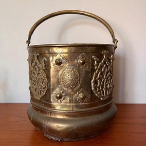 Può includere: Un grande secchio di ottone decorato con un manico. Il secchio presenta un design decorativo con dettagli intricati e un fondo arrotondato.