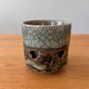 Puede incluir: Una taza pequeña de cerámica gris con un esmalte craquelado y tres recortes en forma de corazón de oro. La taza está sobre una superficie de madera.