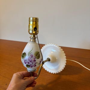 May include: A vintage white glass wall lamp with a painted floral design featuring purple flowers and green leaves. The lamp has a brass-colored metal arm and a white, scalloped shade. The lamp is approximately 8 inches tall.