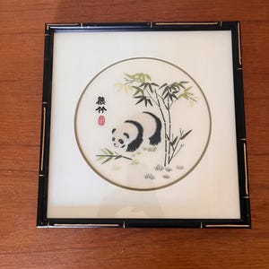 Può includere: Opera d'arte incorniciata con un panda che mangia bambù. Il panda è bianco e nero, su sfondo bianco all'interno di un disegno circolare. La cornice è nera con una texture a bambù. L'opera include caratteri cinesi e un sigillo rosso.