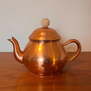 Può includere: Una teiera di rame con un pomo di legno sul coperchio. La teiera ha un corpo arrotondato e un beccuccio laterale. La teiera è appoggiata su una superficie di legno.