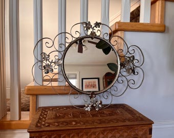 Op de afbeelding: Een zilveren metalen spiegel met een decoratief, gerold frame en kristallen accenten. Het frame is gevormd als een bloem met bladeren en wijnstokken. De spiegel hangt aan een muur.