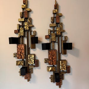 May include: Pair of mid-century modern wall sconces. Each sconce features a geometric design with rectangular and square shapes in gold, copper, and bronze tones. Black candle holders are integrated into the design.