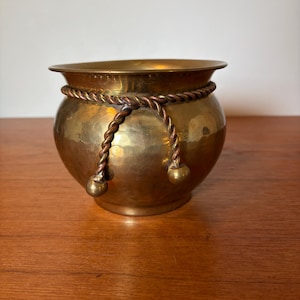 May include: A round, hammered brass bowl featuring a wide, flared rim and a decorative twisted rope detail made of copper-toned metal that encircles the upper body, ending in two small, rounded finials hanging down the front of the vessel.
