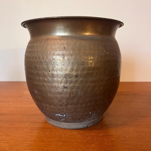 May include: A hammered copper planter with a rounded body and a slightly flared rim. The surface has a textured, dimpled pattern. The planter is a warm, metallic brown colour and sits on a wooden surface.