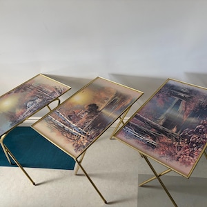 May include: A set of three rectangular TV tray tables with gold-toned metal folding legs. Each tabletop features a printed landscape scene depicting a house near a lake, trees, and autumn-coloured foliage under a bright, hazy sunlit sky.