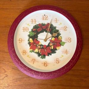 May include: A round wall clock with a dark red crackle-finish frame. The white face features gold numbers, gold hands, and a central Christmas wreath with red poinsettias, yellow ribbons, and small cherubs. Christmas song titles are printed.