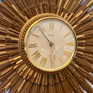 May include: A gold-colored, sunburst-style wall clock. The clock face is cream-colored with gold Roman numerals and hands. The clock's outer frame has a textured, radiating design. The brand name "SYROCO" is printed on the clock face.