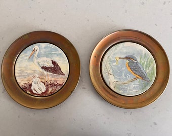 May include: Two brass coasters with a white ceramic insert. One coaster features a white stork with two chicks, the other features a blue kingfisher with a fish in its beak. Both coasters have a brown and white floral design.