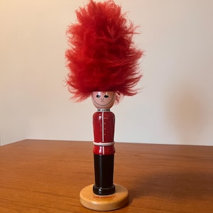 May include: A wooden toy soldier figurine with a red fuzzy hat, red jacket, black pants, and a round wooden base. The face is painted with a smiling expression. The figurine is a decorative item.