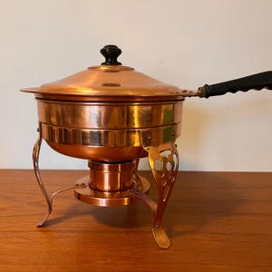 Puede incluir: Un plato calentador de cobre con un asa negra y una tapa. El plato está sobre un soporte con patas decorativas.