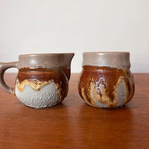 May include: Two handmade ceramic pieces: a small pitcher with a handle and a small jar. Both have a brown, tan, and white glaze. The pitcher has a spout. The jar is round. The pieces are on a wooden surface.