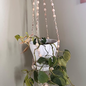 Op de afbeelding: Een witte keramische hangende plantenbak met een hangende groene plant. De plantenbak wordt opgehangen door een macramé hanger versierd met schelpen. De plant heeft hartvormige bladeren met groene en gele variaties.
