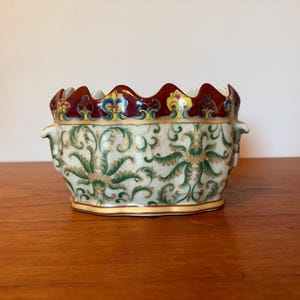 May include: An oval ceramic bowl featuring a scalloped rim with a deep red border and gold trim. The body displays a cream-coloured crackle glaze decorated with intricate green and gold scrolling foliage patterns and small side handles.
