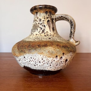 May include: A brown and white ceramic vase with a textured surface and a handle. The vase has a wide base and a narrow neck.
