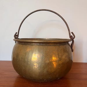May include: A vintage brass pot with a rounded body and a wire handle. The pot has a worn, aged look and is perfect for a rustic or farmhouse decor.