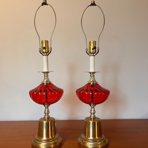 Puede incluir: Par de lámparas de mesa vintage. Cada lámpara presenta una pantalla de vidrio rojo con un diseño acanalado, una base de latón y una columna blanca. Las lámparas tienen una estética moderna clásica de mediados de siglo.