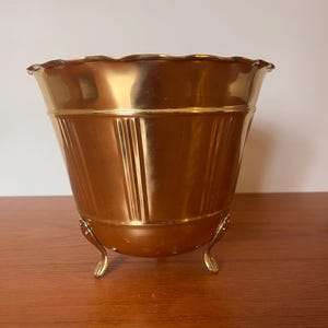 Peut inclure: Un pot de fleur en métal doré avec un bord festonné et des lignes verticales. Le pot a trois pieds incurvés. L'intérieur est de couleur or brillant. Le pot est posé sur une surface en bois.