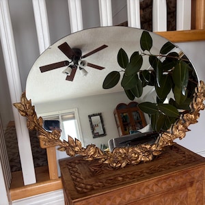 May include: A circular mirror with a gold floral frame. The mirror is placed on a wooden chest. The mirror reflects a room with a ceiling fan and a plant.