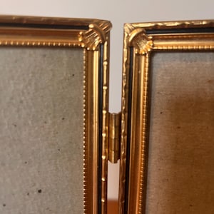 Könnte beinhalten: Ein goldfarbener Bilderrahmen mit schwarzem Innenrand. Der Rahmen ist in der Mitte angeschlagen und hat an den Ecken ein dekoratives Design.