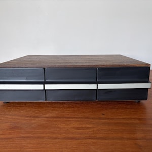 May include: A vintage storage box with a dark wood top and three black drawers, featuring silver-coloured trim. The box is rectangular and rests on a wooden surface. The top has a dark wood grain pattern.