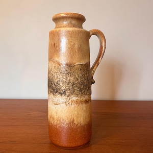 May include: A tall ceramic jug with a handle, displaying a gradient of colours from dark brown to light tan, and a reddish-brown base. The vintage-style jug is likely for decorative use or serving drinks. It measures approximately 30 cm in height.