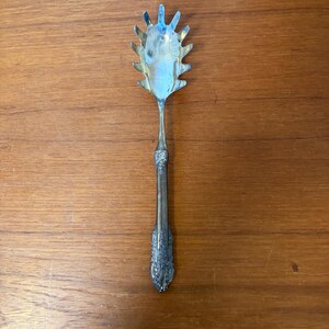 Könnte beinhalten: Ein Vintage-Spaghettilöffel aus Silber mit einem dekorativen Griff und einem geschlitzten, fächerförmigen Kopf. Der Löffel besteht aus glänzendem, reflektierendem Metall und liegt auf einer warmen Holzoberfläche.