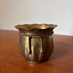 May include: A small, antique brass planter with a scalloped rim and a decorative rope detail around the middle. The planter has a rounded, ribbed body and sits on a wooden surface. The brass has a warm, aged patina.