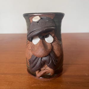 May include: A handmade ceramic mug featuring a sculpted face of a man with a hat, mustache, and beard. The mug is mainly brown with dark green and white accents. The mug is on a wooden surface.