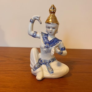 May include: A white porcelain figurine of a woman in a blue and gold costume, sitting with her right arm raised and her left hand holding a fan. The figurine is decorated with gold accents and has a blue and gold headdress.