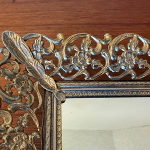 May include: A rectangular vanity tray featuring an ornate, metallic gold-toned filigree border with intricate floral and scrollwork patterns. The frame has a raised, decorative edge with a textured finish, surrounding a central reflective mirror surface.