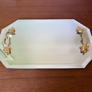 Puede incluir: Una bandeja de espejo octogonal con una superficie de vidrio reflectante y bordes biselados. La bandeja cuenta con dos asas de metal dorado ornamentadas fijadas en los lados opuestos, cada una con detalles intrincados en forma de hojas.