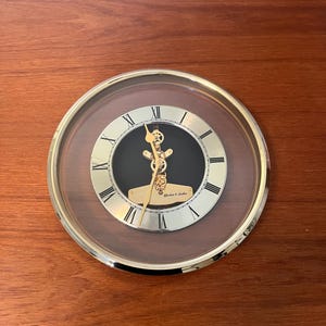 May include: A round wall clock with a gold-colored bezel and a clear face. The clock face is black with gold Roman numerals and hands. The clock has a decorative gold pendulum and the text "Gordon C. Lindes".